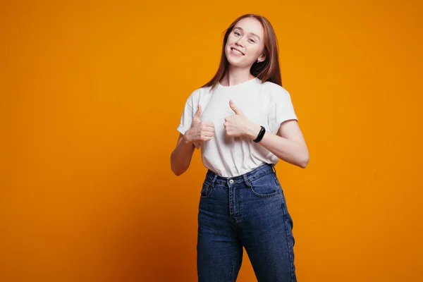白いTシャツを着た赤い髪の女の子は、承認の印として親指を持ち、オレンジの背景に正しい選択. — ストック写真