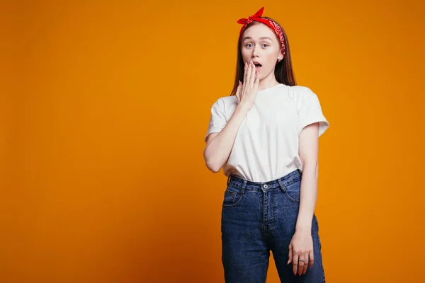 Redhead girl with a bandana on her head in white T-shirt surprised covered mouth with her hand on an orange background in studio.