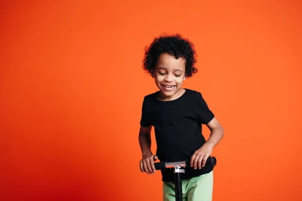 Pequeno menino etíope de cabelos encaracolados em uma camiseta escura parece envergonhado e sorri enquanto monta uma scooter no estúdio — Fotografia de Stock