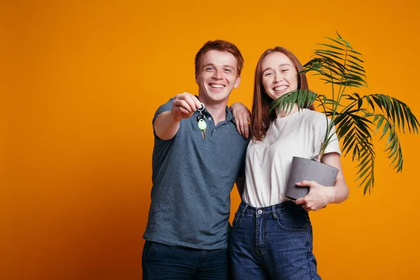 Рыжеволосый парень берет свою жену с зеленым растением и протягивает ключи от новой квартиры в камере в студии на оранжевом фоне — стоковое фото