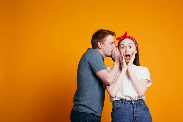Рыжий парень шепчет на ухо своей девушке, и она очень удивлена неожиданностью. Эмоциональная студия на оранжевом фоне — стоковое фото