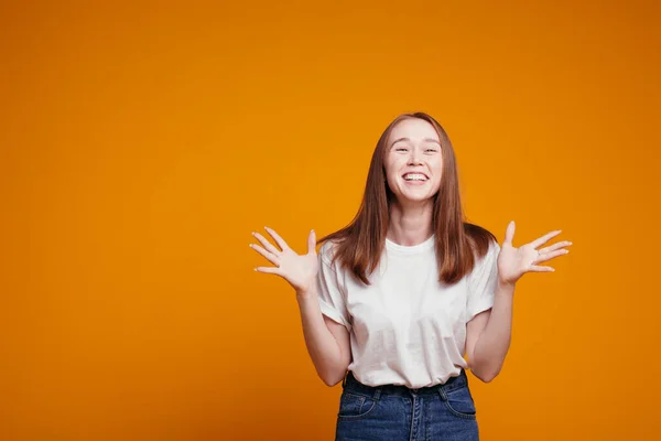 Fille aux cheveux roux dans un T-shirt blanc gesticule avec ses mains et rit de toute sa bouche se réjouissant de réductions ou une surprise soudaine sur un fond orange — Photo