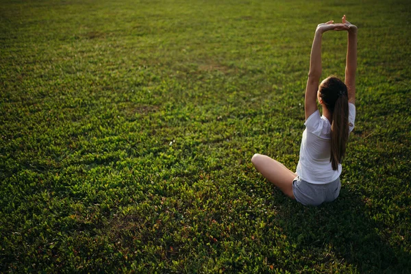 Top view of yoga trainer stretching arms up while sitting in lotus position while meditating in yoga on the lawn at sunset.