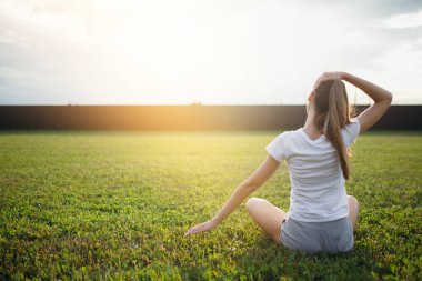 Yeşil bir çimenlikte oturan ve lotus pozisyonunda otururken boynunda egzersiz yapan güzel bir kızın manzarası..