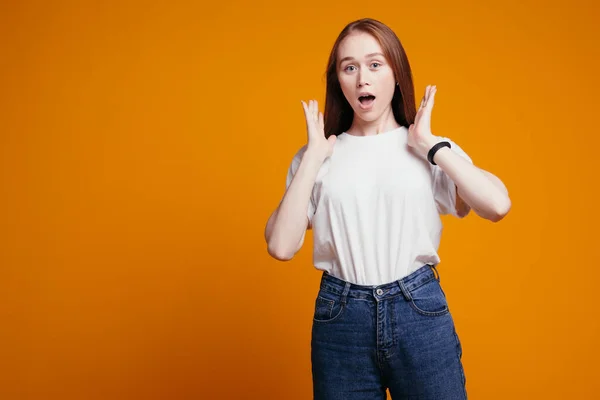 La fille aux cheveux roux tient étonnamment ses mains près de son visage et ouvre la bouche avec surprise sur un fond orange en studio. — Photo