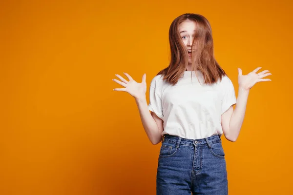 Crazy red-haired girl bounces with joy and spreads her arms to the sides, hair covers her face. Orange background.