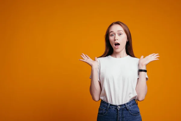 Portret van een roodharig Europees meisje dat haar armen naar de zijkant spreidt en naar de zijkant kijkt en niet weet wat te kiezen. Oranje achtergrond. — Stockfoto