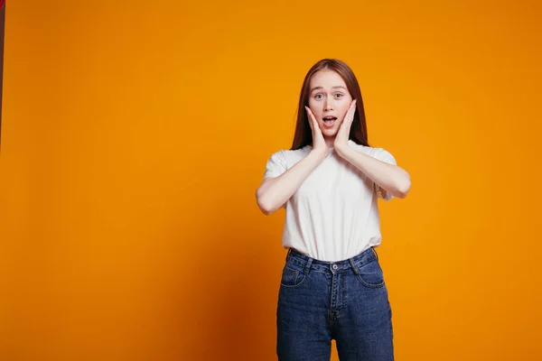 La fille aux cheveux roux en T-shirt blanc tient ses mains sur son visage dans la surprise et ouvre la bouche dans la surprise. Prise de vue émotionnelle sur fond orange. — Photo