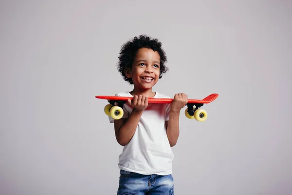 Curly menino africano pressiona um longboard vermelho para si mesmo e sorri para a câmera no estúdio em um fundo leve — Fotografia de Stock