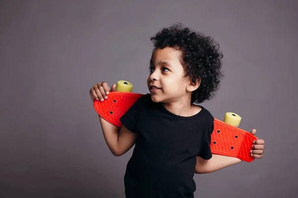 Close-up retrato de menino afro-americano em uma camiseta preta segurando skate vermelho atrás de suas costas e sorrindo . — Fotografia de Stock