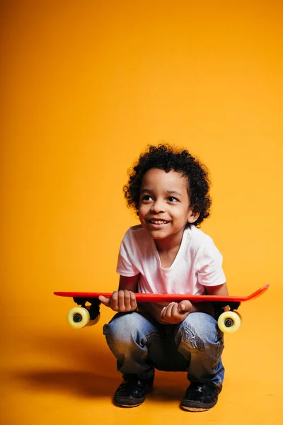 Donkerhuid krullend donker gevild jongen hurkt en houdt op zijn schoot een rood longboard en lacht op een gele achtergrond — Stockfoto
