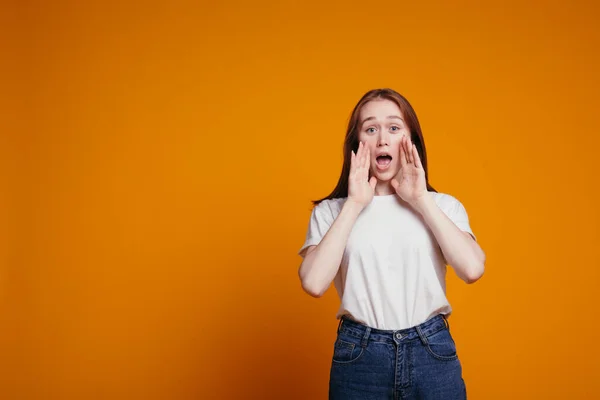 Retrato de uma menina de cabelos vermelhos em uma camiseta branca apertou as mãos em sua boca e gritando alto. Tiro emocional em um fundo laranja . — Fotografia de Stock