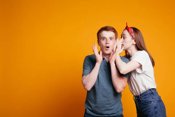 Una chica pelirroja linda susurra una sorpresa en su oído novios y él está muy sorprendido. Disparo emocional sobre un fondo naranja — Foto de Stock