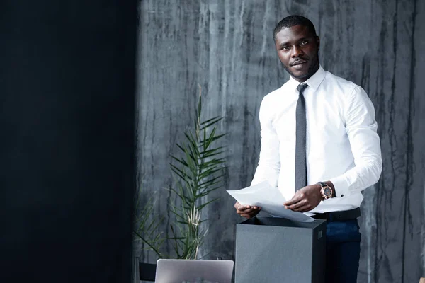 Czarny mężczyzna w białej koszuli stojący przed biurkiem, trzymający dokument i patrzący w kamerę — Zdjęcie stockowe