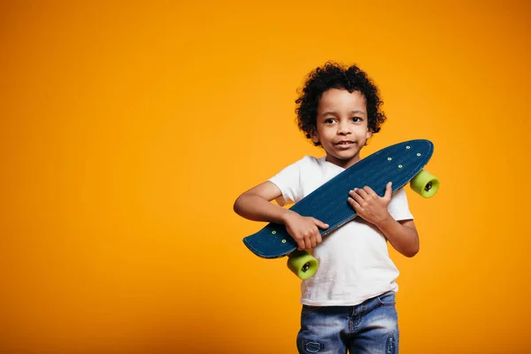 African American Boy en T-shirt blanc et jeans étreint une planche à roulettes et la presse sur sa poitrine sur fond orange. — Photo