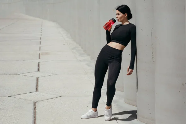 Linda menina asiática esportiva se inclina na rua da parede depois de um longo treino e água potável de uma garrafa vermelha . — Fotografia de Stock