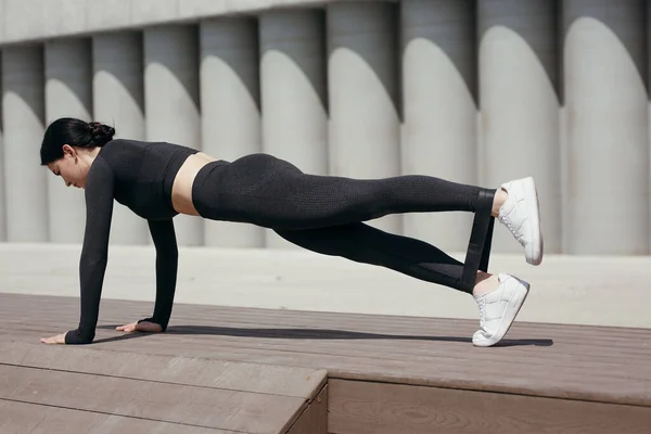 Fata antrenor de fitness este într-o poziție și scândură face exerciții cu o bandă de cauciuc sport la gleznele de pe picioarele de pe strada din parc — Fotografie, imagine de stoc