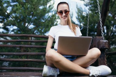 Güneş gözlüklü bir kız bacak bacak üstüne atmış bir bankta oturuyor ve elinde dizüstü bilgisayarla oturuyor. Şehir dışında iş ve açık hava aktiviteleri.