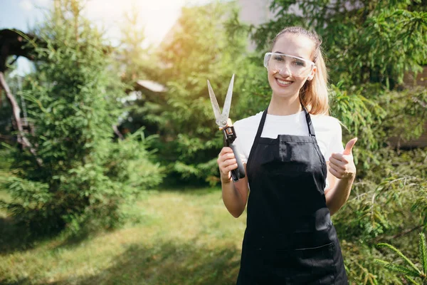 Chica jardinero atractiva en un delantal y gafas de seguridad sostiene tijeras de jardín en sus manos y muestra el pulgar hacia arriba sobre un fondo de vegetación verde — Foto de Stock