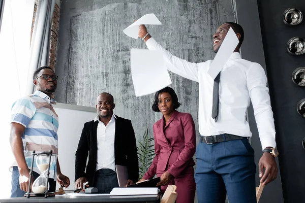 Ein schwarzer Afroamerikaner im weißen Hemd wirft Papiere mit einem Vertrag, und der Rest des Personals lacht und freut sich über den Deal im Büro. — Stockfoto