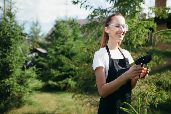 Chica morena cuidando plantas. Trabaja en el invernadero. Tijeras arbustos y árboles con tijeras de hierba — Foto de Stock