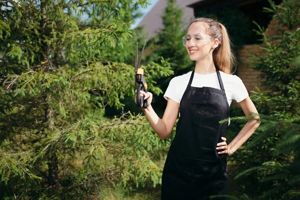 Retrato de una niña jardinero sobre un fondo de árboles en una casa de campo de verano. Latina se para con cortadores de pincel en sus manos y sonríe — Foto de Stock