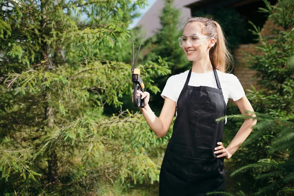 Chica jardinero se encuentra en un delantal negro con tijeras para la hierba en sus manos y sonrías.Trabajar en la primavera en la parcela nature.garden — Foto de Stock