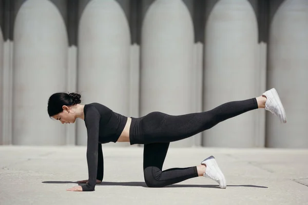 Fitness edző egy szűk testalkatú melegítőben négykézláb áll és felemeli a lábát, hogy felpumpálja a comb hátsó izmait a parkban a szabadban. — Stock Fotó