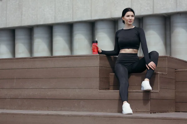 Deportes chica asiática en traje negro sentado en un podio de madera en la calle y descansando después del entrenamiento con una botella de agua en la mano —  Fotos de Stock