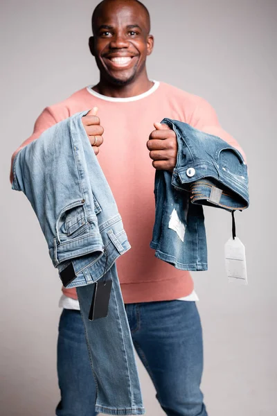 Uśmiechnięty, ciemnoskóry Afroamerykanin trzyma dżinsy nie wiedząc, co wybrać. Badanie sklepu odzieżowego — Zdjęcie stockowe