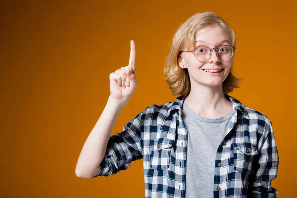 Милая девушка в клетчатой рубашке и очках смотрит вверх и показывает пальцем вверх на текст на оранжевом фоне — стоковое фото