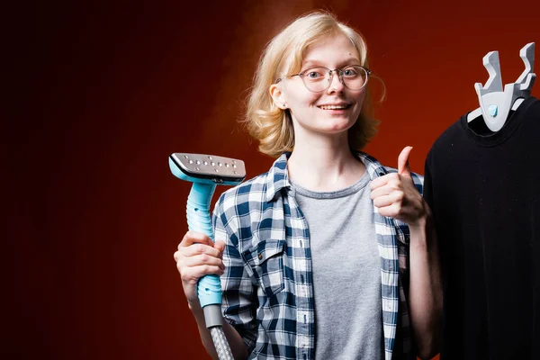 Chica feliz con el pelo rubio sosteniendo un vapor en la mano y muestra el pulgar. Limpieza química de los trabajadores sobre un fondo rojo . —  Fotos de Stock