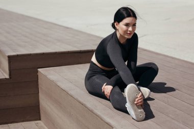 Pilates antrenörü ısınma egzersizleri yapıyor. Parktaki sokakta tahta bir zeminde otururken bacağa yaslanıyor.