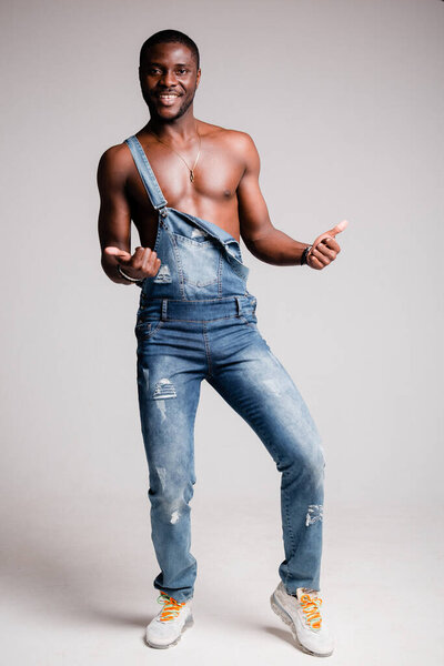 Dark-skinned African-American man with naked torso in denim pants with suspenders crossed his arms on his chest