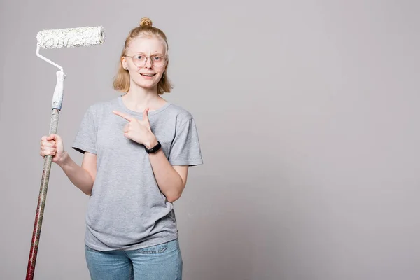 Atractivo pintor niña sosteniendo un rodillo profesional de la construcción. La chica muestra el rodillo con tu dedo índice . —  Fotos de Stock