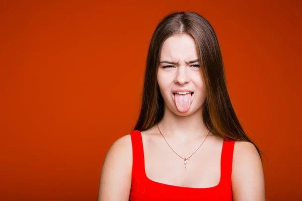 Emocjonalny portret młodej dziewczyny z długimi włosami, która przymrużyła oczy i pokazuje język przed kamerą na pomarańczowym tle — Zdjęcie stockowe