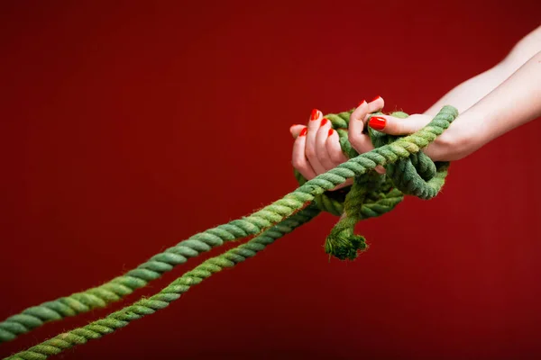 Female hands are tangled in a green rope on a burgundy background. The concept of problems that pull down and the inability to get rid of them