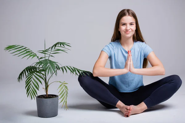 A girl sits on a white background and meditates in a lotus position while sitting next to a green flower. The concept of meditation and female yoga — Stock Photo, Image