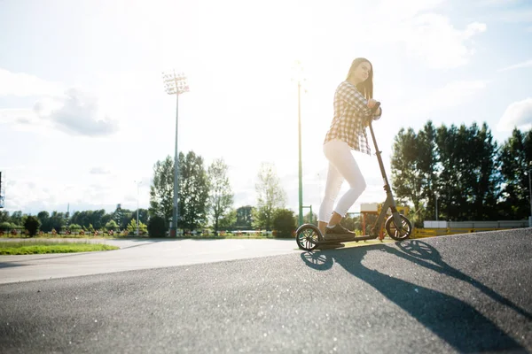 Retrato callejero de una adolescente con una camisa a cuadros montada en un scooter en un parque sobre un fondo al atardecer. Publicidad rodaje de artículos deportivos —  Fotos de Stock
