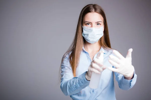 Trabalhadora de escritório menina fica em uma máscara médica e polvilha um anti-séptico em luvas, a fim de prevenir a doença por doenças em um fundo cinza — Fotografia de Stock