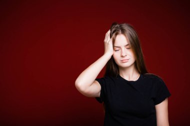 Kırmızı arka plandaki baş ağrısı yüzünden elini başının üstünde tutan siyah tişörtlü bir kızın portresi.