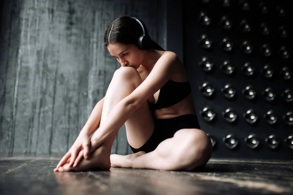 Chica en ropa atlética negra para ejercicios de yoga para estirar los músculos de las manos y escuchar música a través de auriculares . — Foto de Stock
