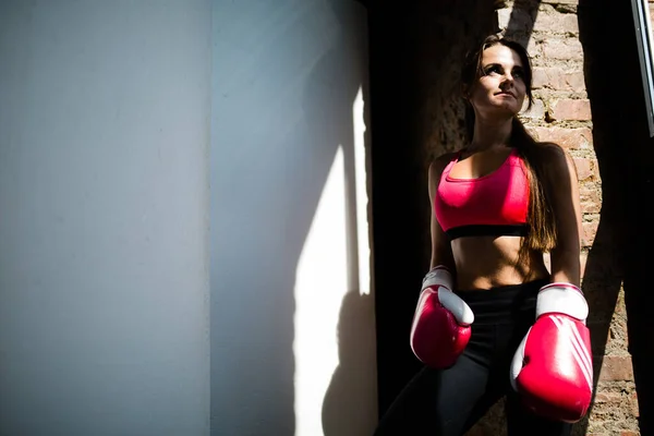 A atleta menina em luvas de boxe vermelho fica perto da janela de vidro e olha para o amanhecer após o treinamento matutino . — Fotografia de Stock