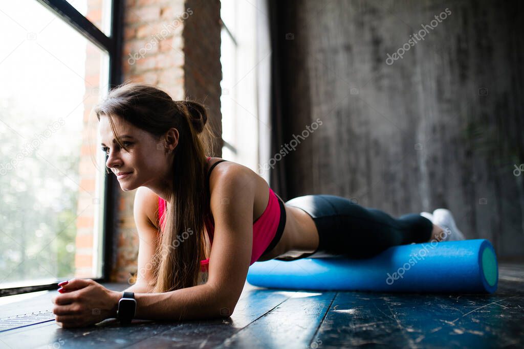 A girl does a plank exercise using a sports cylinder to work through the fascia of the femoral muscles near panoramic window.