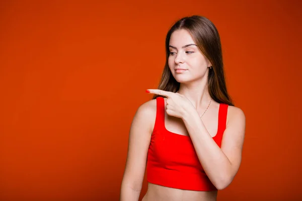 A menina com cabelos longos em uma camisa vermelha mostra o dedo indicador para o lado em um fundo laranja — Fotografia de Stock
