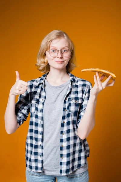 Švédská dívka s brýlemi a kostkovanou košilí ukazuje prst jako a drží plátek pizzy v rukou na oranžovém pozadí — Stock fotografie