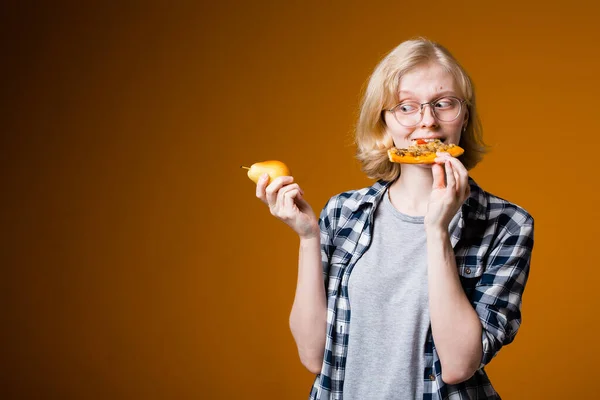 Het meisje dat blond is bijt een stuk pizza af met een peer in de andere hand. Junk en gezond voedsel concept — Stockfoto