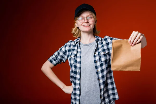 Κορίτσι υπάλληλος ενός εστιατορίου fast food κατέχει μια έτοιμη παραγγελία σε μια χάρτινη σακούλα και δείχνει τον αντίχειρα στον πελάτη. — Φωτογραφία Αρχείου