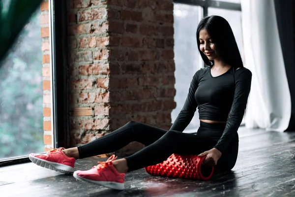 Una entrenadora de Pilates en un chándal negro se sienta en el piso del gimnasio y pone un rollo de masaje debajo de su muslo para ejercitar los músculos de la fascia. . — Foto de Stock