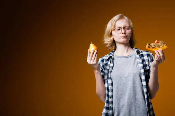 A menina loira de óculos segura uma pêra e uma fatia de pizza em suas mãos pensando o que comer. O fim de uma dieta saudável e não saudável em um fundo laranja . — Fotografia de Stock